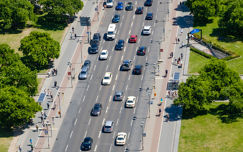 Straßenszene mit verschiedenen umweltfreundlichen Verkehrsmitteln: Elektroautos, E-Bikes, Straßenbahn und Fußgänger.