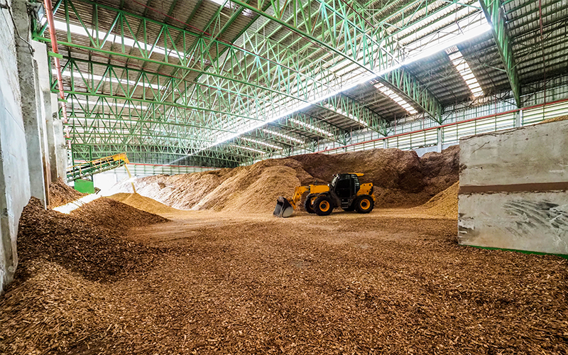 Das Bild zeigt eine Industriehalle, in der Holzhackschnitzel mit einem Bagger bewegt werden.