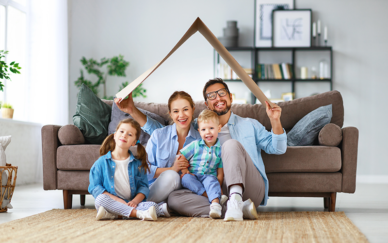 Familie mit angedeutetem Dach aus Pappe, sitzt im Wohnzimmer auf dem Boden