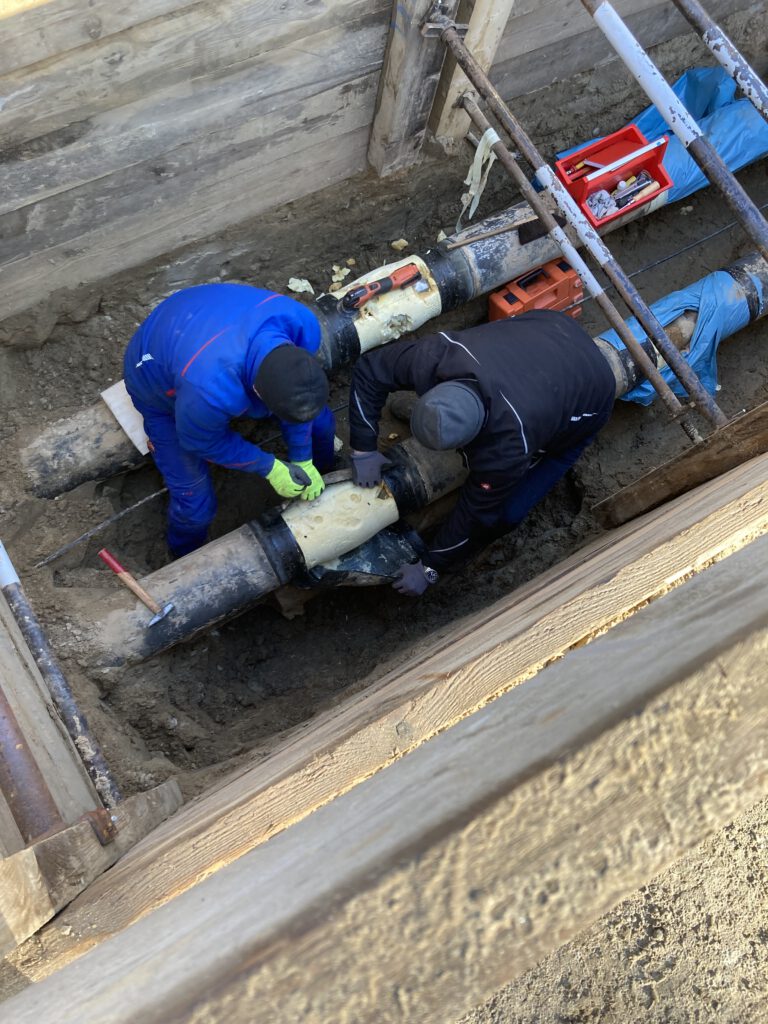 Zwei Bauarbeiter arbeiten in einer Baugrube an einer großen Rohrleitung. Sie dämmen ein schwarzes Rohr mit hellem Isoliermaterial.
