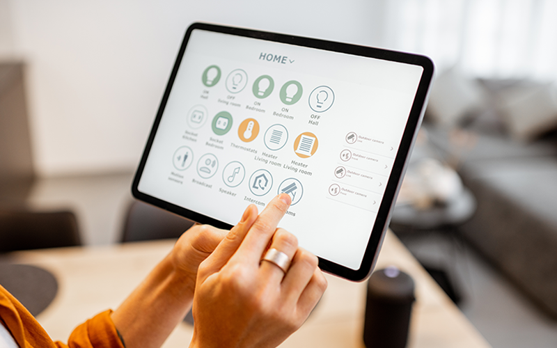 Hand hält Tablet mit Smart-Home-Steuerungs-App und tippt auf Display mit Icons für verschiedene Geräte.