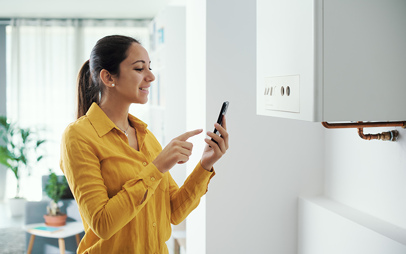 Frau mit gelber Bluse bedient Smartphone vor weißer Wand mit Warmwasser-Boiler.