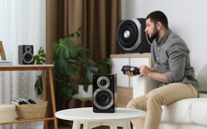 Lautsprecher stehen auf einem runden weißen Tisch vor einem Mann, der eine Fernbedienung hält. Im Wohnzimmer sind weitere Lautsprecher und Pflanzen zu sehen.