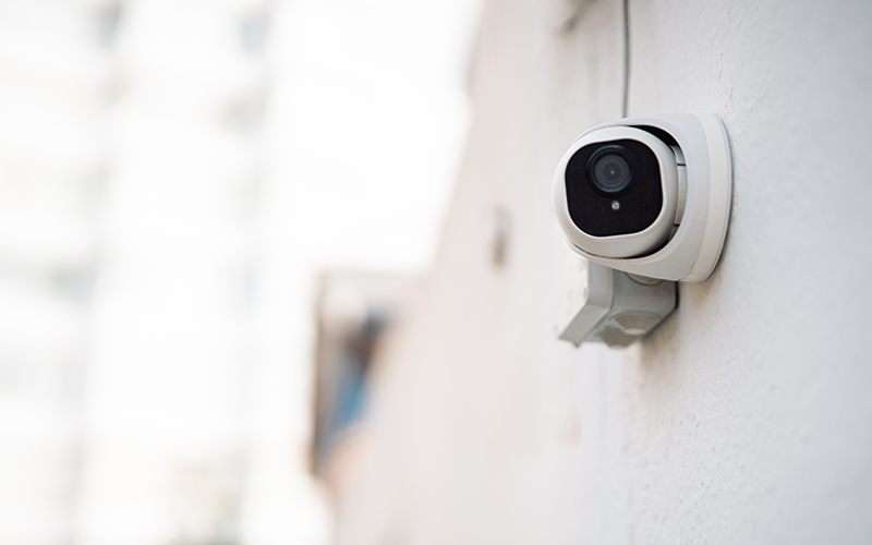 Weiße Überwachungskamera mit schwarzer Linse an einer Außenwand montiert.