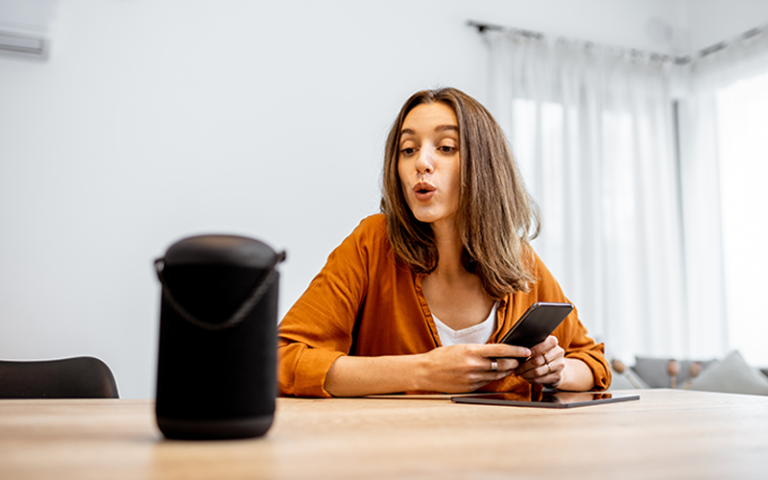 Frau mit orangefarbener Bluse sitzt am Tisch und hält Smartphone. Vor ihr steht ein schwarzer Sprachassistent.
