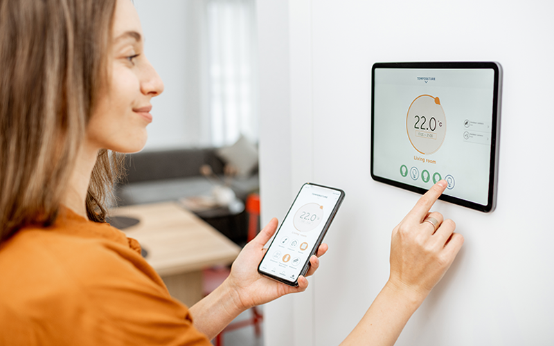 Person bedient an der Wand montiertes Smart-Thermostat mit Touchscreen und hält Smartphone mit passender App in der Hand.