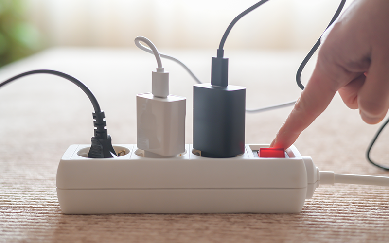 Hand drückt roten Schalter an weißer Steckdosenleiste mit mehreren eingesteckten Netzteilen auf Holztisch.