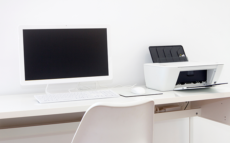 Weißer Schreibtisch mit weißem Computerbildschirm, Tastatur, Maus und weißem Drucker vor weißer Wand.