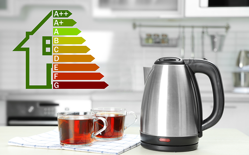 Elektrischer Wasserkocher aus Edelstahl neben zwei Glastassen mit Tee vor einer Küchenarbeitsplatte und einem Energieeffizienzlabel in Form eines Hauses mit den Klassen A++ bis G.