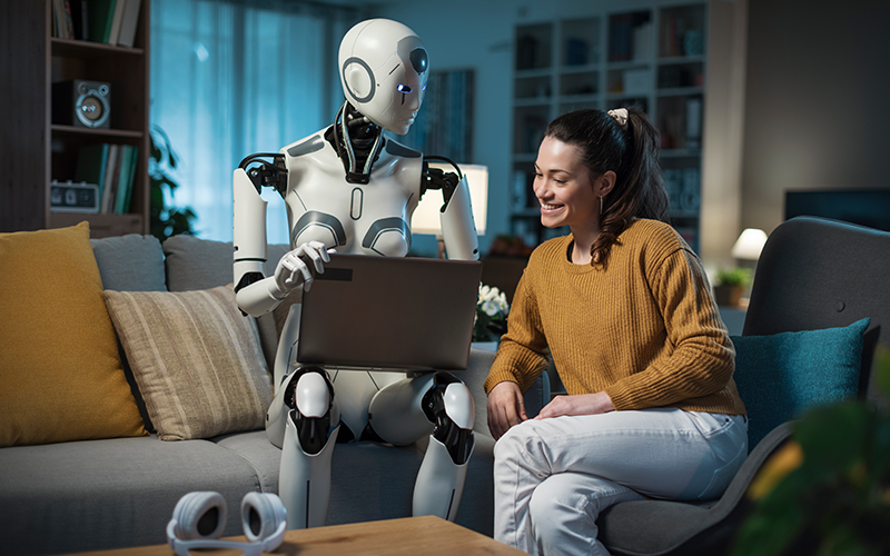 Roboter mit menschlichem Design hält Laptop auf einem Sofa neben sitzender Person in senfgelbem Pullover.