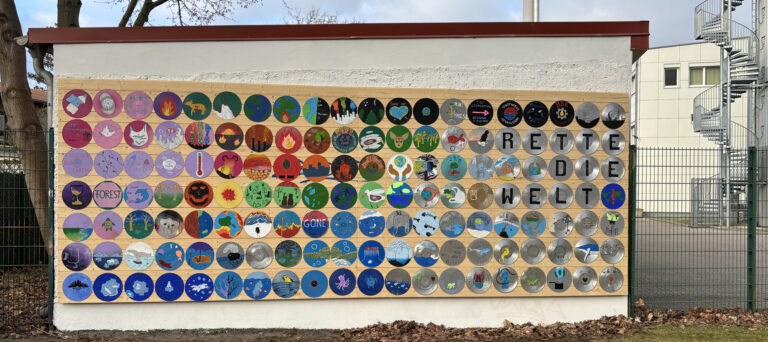 Viele bunte Kreise mit gemalten Bildern hängen an einer Wand. Die Bilder zeigen Themen wie Umwelt, Natur und Klima. Auf der rechten Seite steht: „Rette die Welt“.