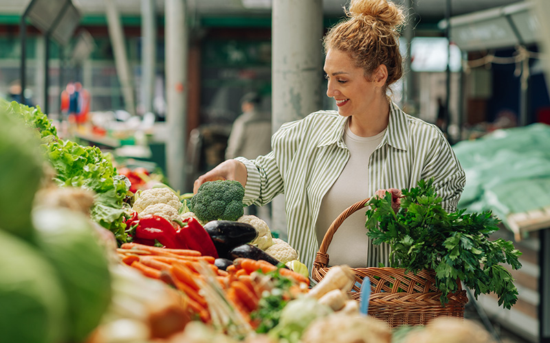 Frau mit Korb wählt frisches Gemüse an einem Marktstand.