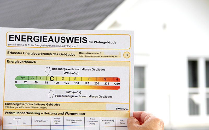 Hand hält Energieausweis für Wohngebäude mit farbiger Skala von A+ bis H zur Einordnung des Energieverbrauchs.