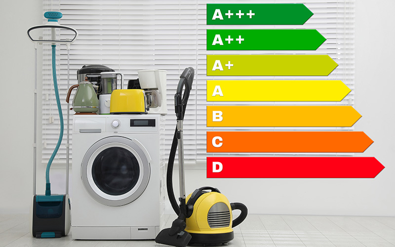 Waschmaschine, Staubsauger, Wasserkocher und Toaster stehen nebeneinander vor einer weißen Wand mit einem farbigen Energieeffizienzlabel mit den Pfeilen von A+++ bis D.