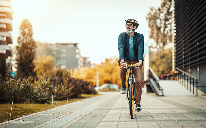 Mann mit Helm fährt Fahrrad auf einem städtischen Radweg neben einem modernen Gebäude und herbstlich gefärbten Bäumen.
