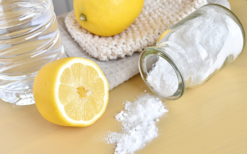 Halbierte Zitrone, ganze Zitrone, Glas mit verschüttetem weißem Pulver, Plastikflasche mit klarem Wasser und zwei gefaltete Handtücher auf hellem Untergrund.