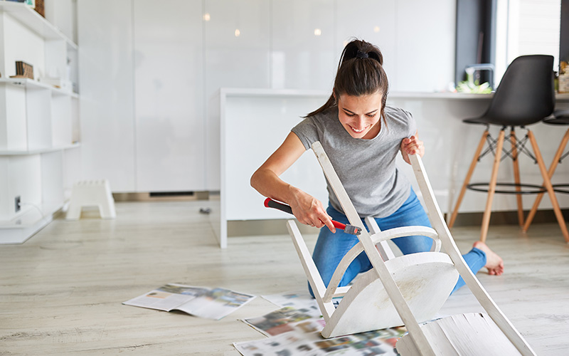 Frau in grauem T-Shirt und blauer Hose bearbeitet einen weißen Holzstuhl, der umgedreht auf dem Boden liegt, mit einem Werkzeug.
