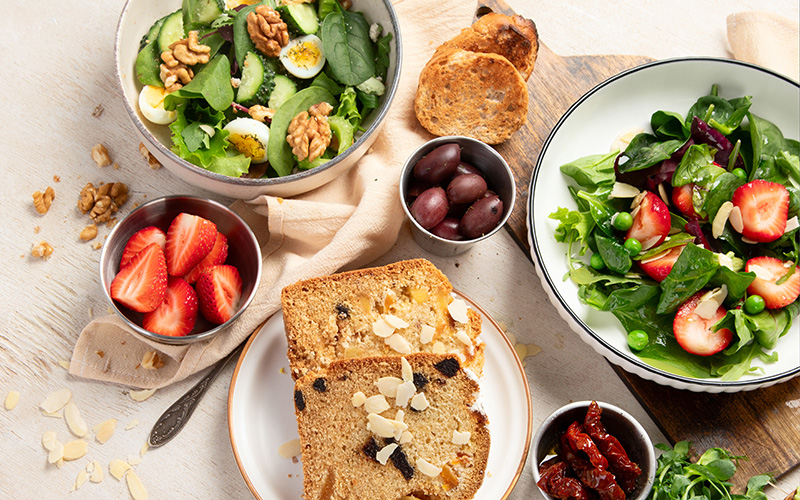 Gefüllte Gefäße und Schalen mit Brot, Salat, Erdbeeren, Oliven und getrocknete Tomaten stehen auf einem hellem Tisch.