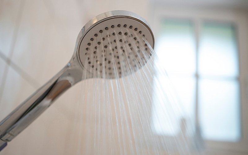 Nahaufnahme eines Duschkopfs, aus dem Wasserstrahlen fließen, im Hintergrund ein Fenster.