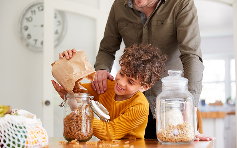 Kind schüttet Nudeln aus einer Papiertüte in ein Glasgefäß, daneben sind ein weitere Glasgefäß mit Getreide und Netzbeutel zu sehen.