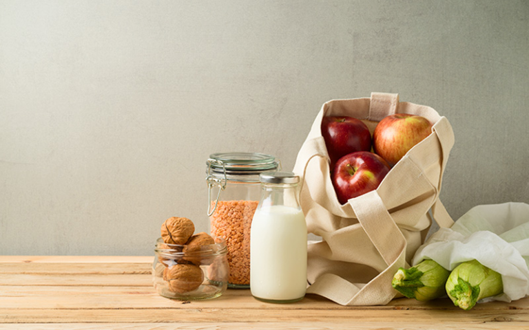 Mehrweg-Stoffbeutel mit Äpfeln und Zucchinis, daneben Glasgefäße mit Walnüssen und roten Linsen sowie eine Glasflasche mit Milch auf einem Holztisch.
